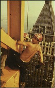 A man in a red hard hat sits on a girder high above New York City