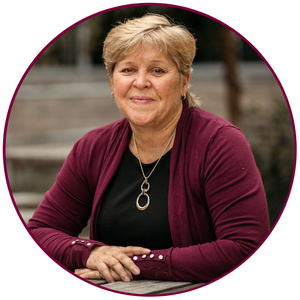 A photo of Heather Sheardown. She is wearing a black shirt and a maroon cardigan. She is seated and resting her elbows and forearms on a surface in front of her. She is smiling directly at the camera.