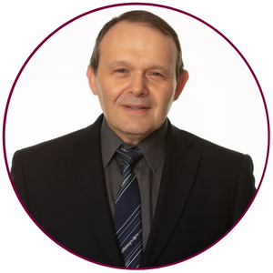 A headshot of Igor Zhitomirsky. He is wearing a grey dress shirt, a dark tie and a black suit jacket. He is looking directly at the camera and is standing against a white backdrop.