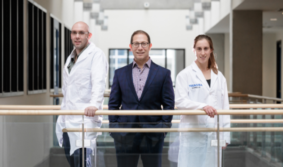 Three researchers, one wearing a business suite and the other two wearing white lab coats, pose for a picture in front of a glass-enclosed walkway