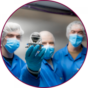 Three people in blue lab coats and hairnets. One of them is holding up the payload of the McMaster NEUDOSE satellite