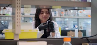 A researcher wearing a white lab coat and black medical gloves holding up and examining a small item 