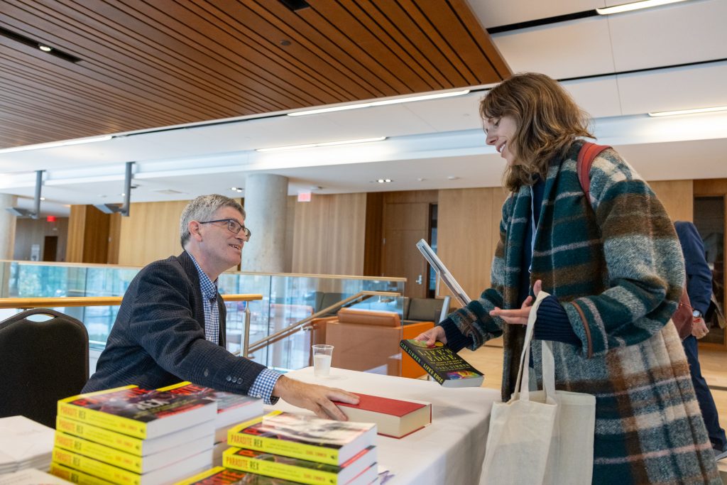 Carl Zimmer in conversation with another person. Zimmer is seated at a table that has his books on it 