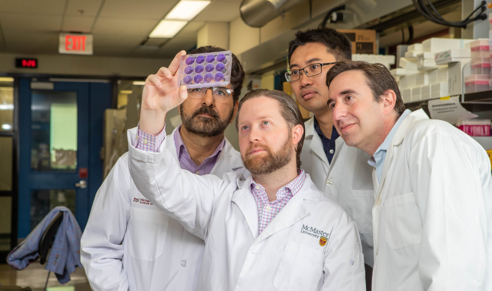 Four men in lab coats standing in a lab looking at samples that one is holding up to the light.