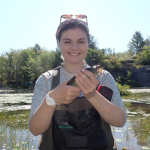 A student holds a fish in front of a lake.