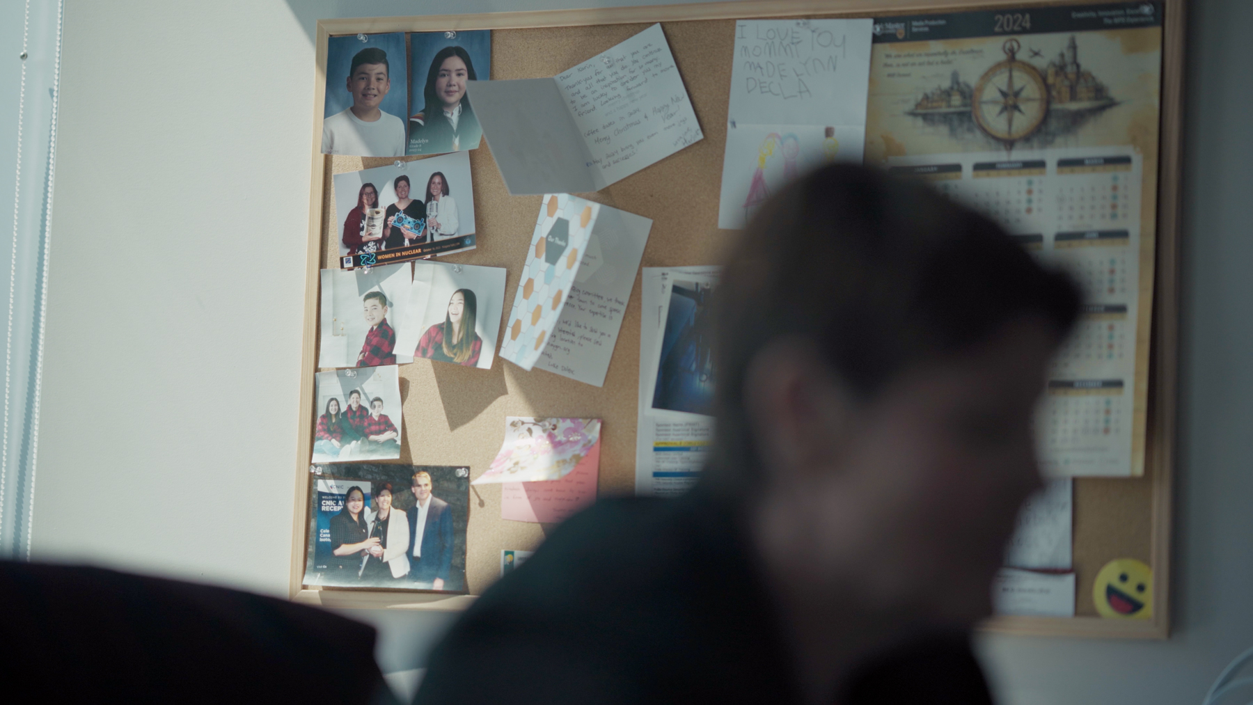 A cork board with a calendar, cards and photos pinned to it. The profile of Karin Stephenson’s face is in the foreground, out of focus. 