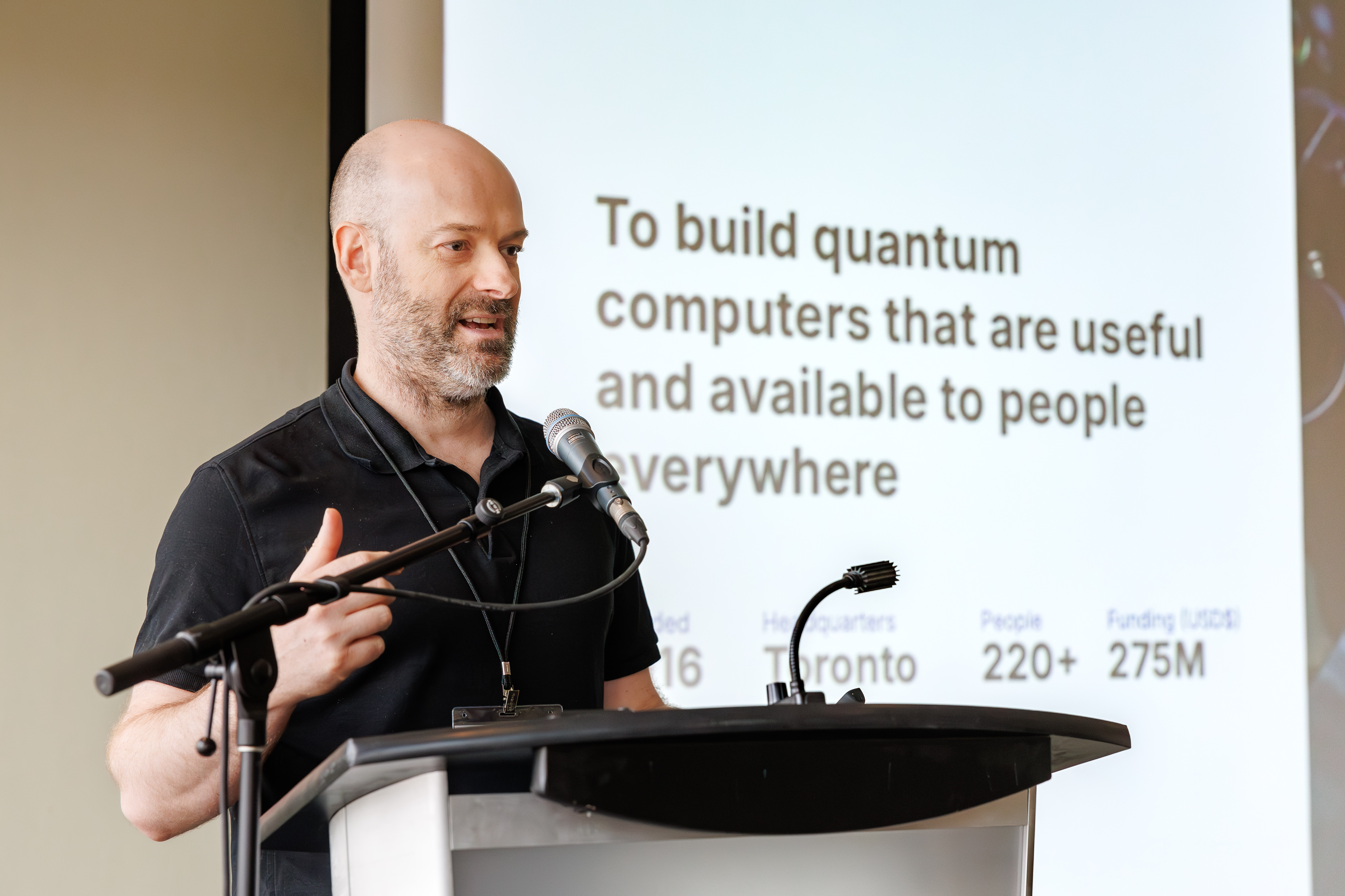 A man in a black shirt speaks into a mic at a podium in front of a slide presentation about building quantum computers.