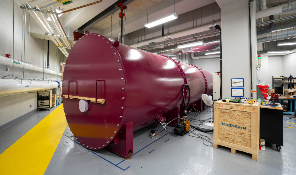 A large, maroon cylindrical neutron beam inside of a room with grey, concrete floors. 