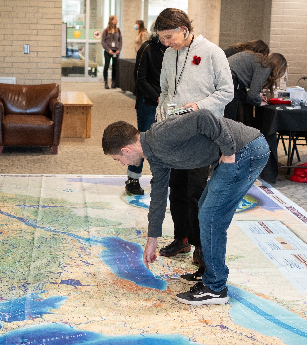 A person in jeans bends down to point to a spot on the enormous floor map. A second person is standing beside him on the map, also looking down at it.