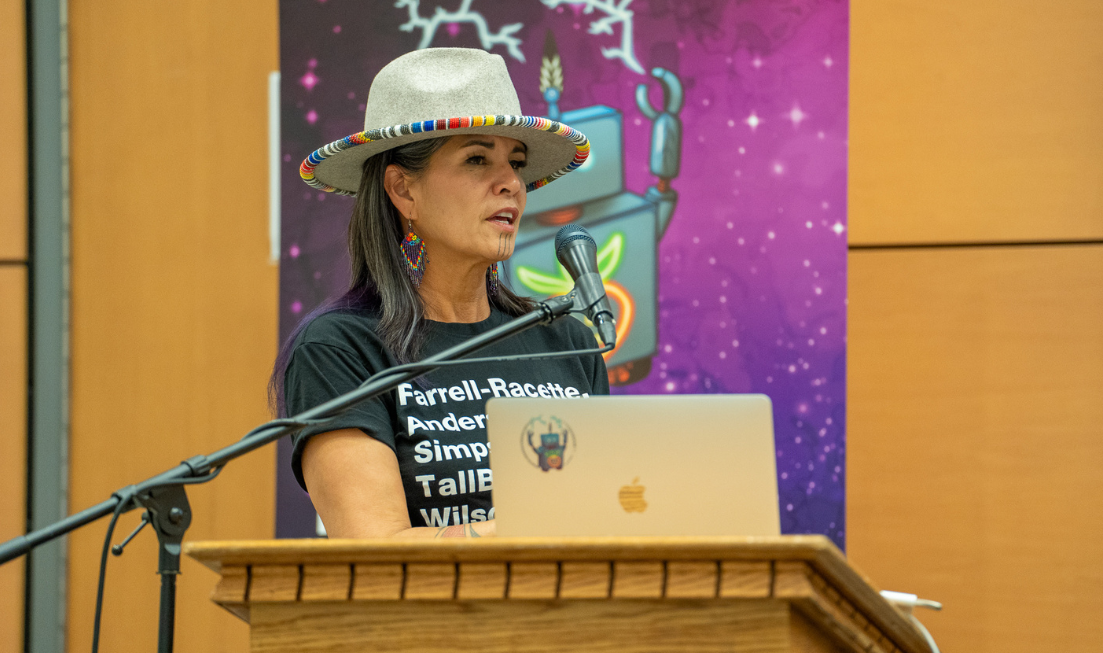 MIRI director Savage Bear, in a hat and a black T shirt, speaks at the podium in front of a MIRI banner.