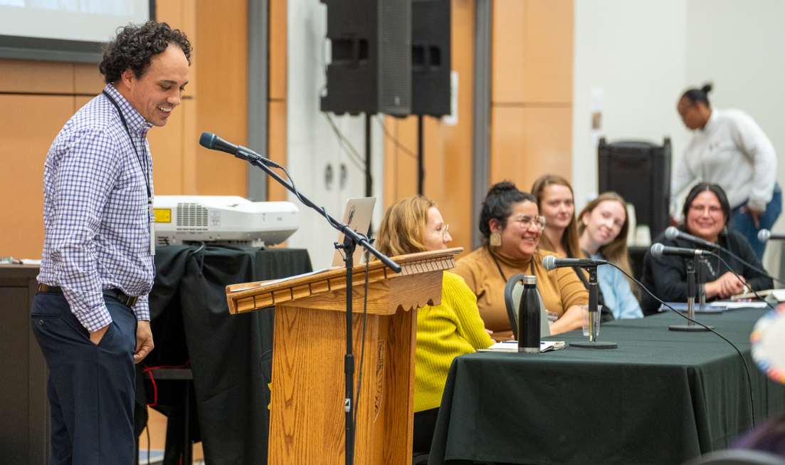 Allan Downey stands at the podium, looks down and smiles with his hand in his pocket. Beside the podiu, a panel of speakers and presenters all smile.