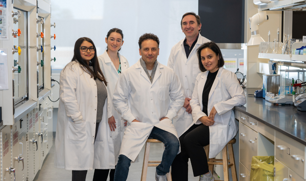 Five researchers all wearing white lab coats and smiling at the camera. They are in a laboratory setting. 