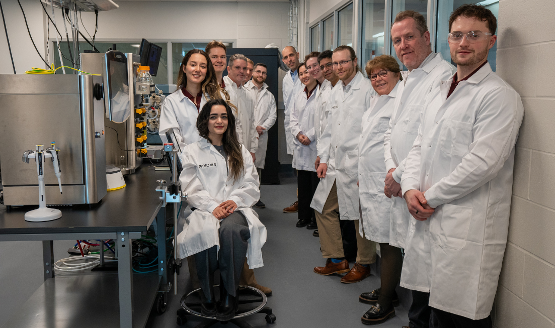 About a dozen smiling people in lab coats line up for a group photo in a lab.