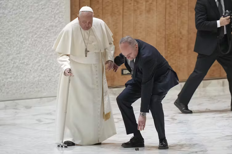 Pope Francis stands, leaning on a cane, while a man in a suit next to him leans down to grab a piece of the cane that has fallen off.