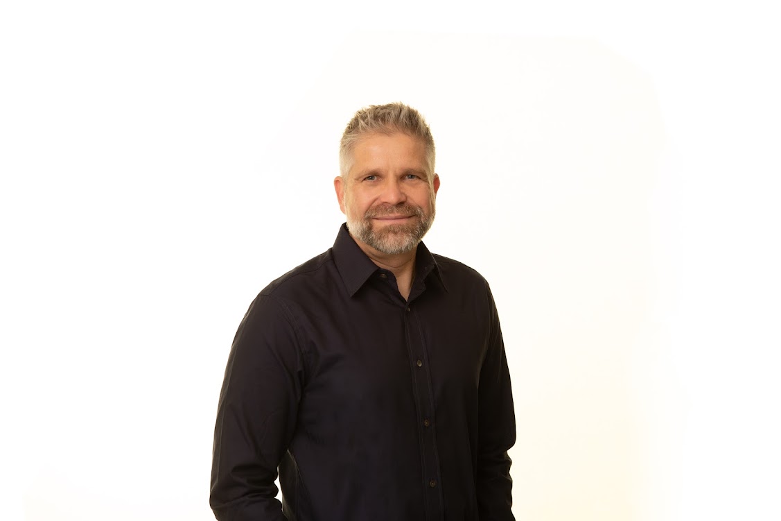 A man in a black dress shirt smiles at the camera.