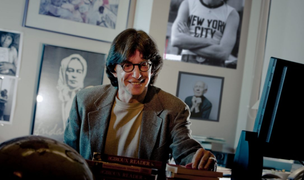 A smiling Henry Giroux standing at a desk with a wall of photos behind him.