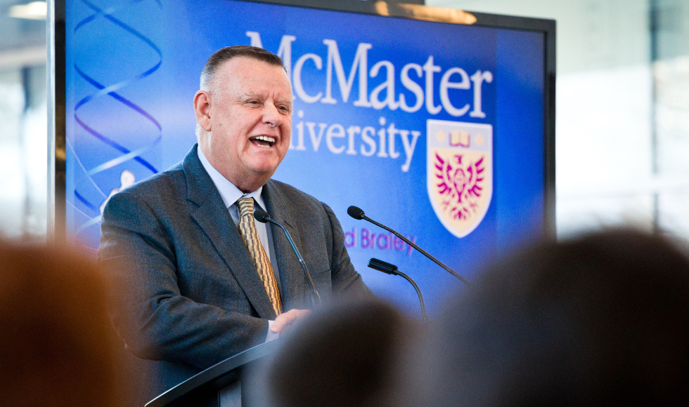 A smiling man speaks at a lectern in front of a banner that says McMaster University