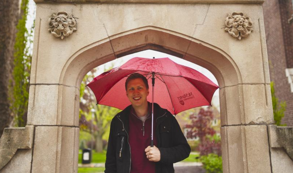 A man stands with a red umbrella under Edwards Arch at McMaster University