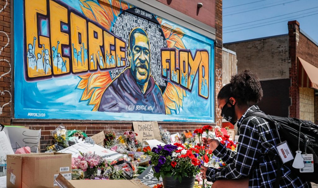 A person adds a bouquet to a growing pile of flowers beneath a painted mural of George Floyd