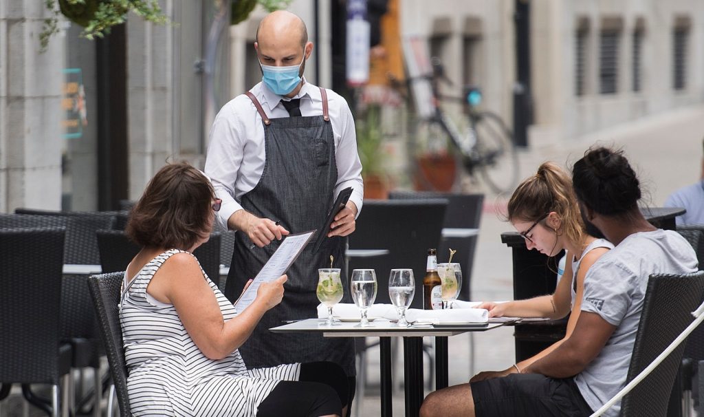 A server wearing a mask talks to a person holding a menu at a table on a patio alongside two others.