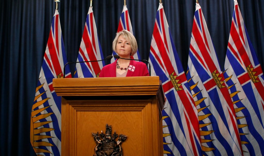 Bonny Henry stands behind a podium under bright lights with British Columbian flags behind her. She doesn't look pleased.