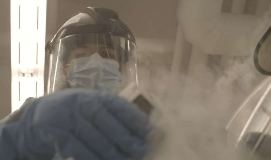 A scientist wearing full protective lab gear, including gloves, face shield, mask and gown, is carefully holding something with smoke or mist coming off it.