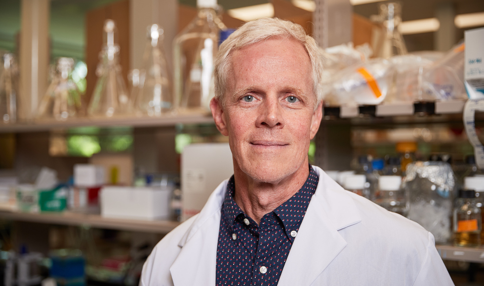 A smiling man stands in front of a lab shelf with beakers on it