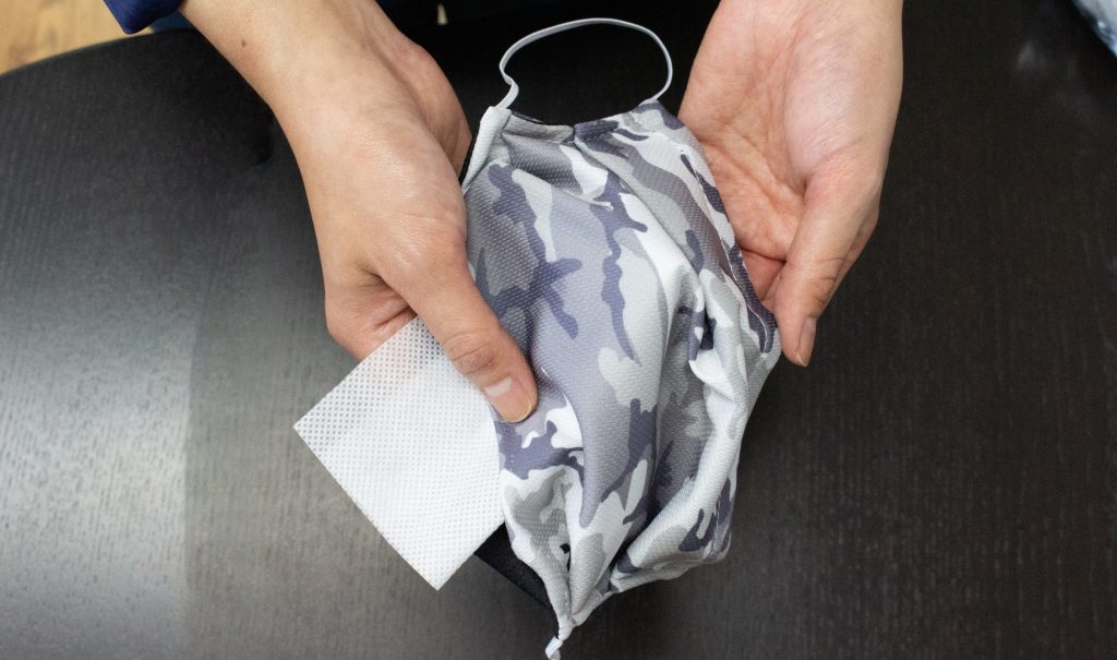 Close-up of hands holding a grey fabric mask, with a square of white polypropylene filter material sticking out from under it.