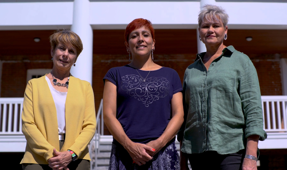 Three women stand in front of a house with a white railing