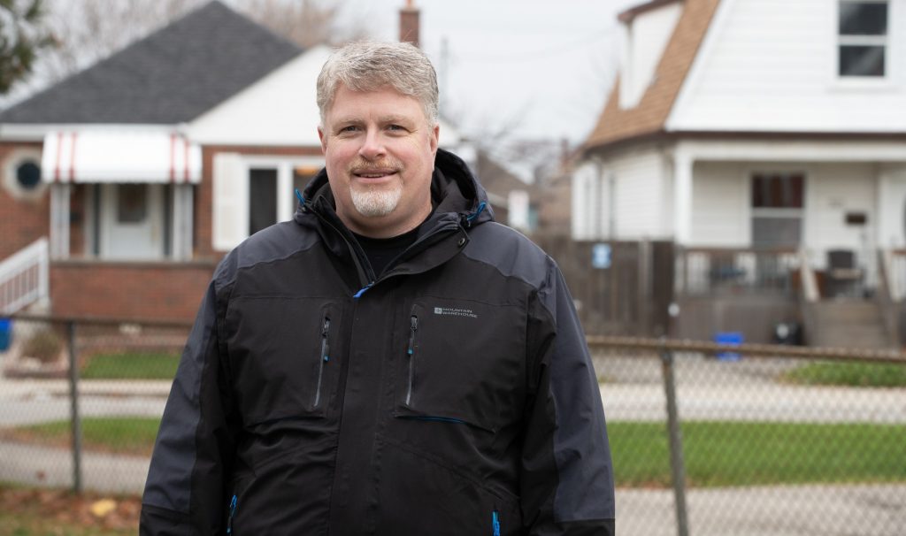 Jim Dunn is smiling at the camera in the foreground. Behind him are small houses on a residential city street.