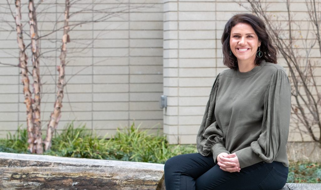 Vanessa Watts sitting on a log bench outside a building, smiling.