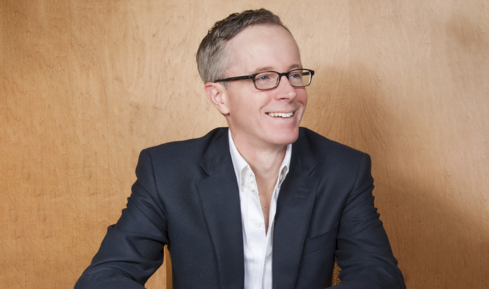 The photo is of a smiling man wearing glasses, a white shirt and a suit jacket