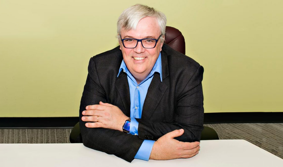 A man in a dark balzer and blue shirt sits at a desk, smiling