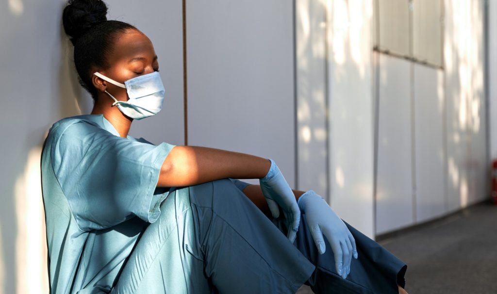 Tired person in scrubs and a mask sitting on the floor.