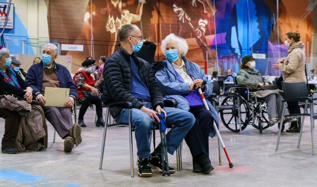 Masked older adults sitting waiting in chairs that are spaced out for distancing.