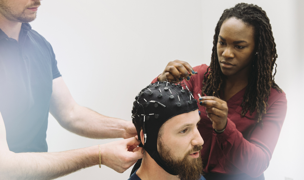 A person is seated while two people attach electrons to a cap sitting on his head