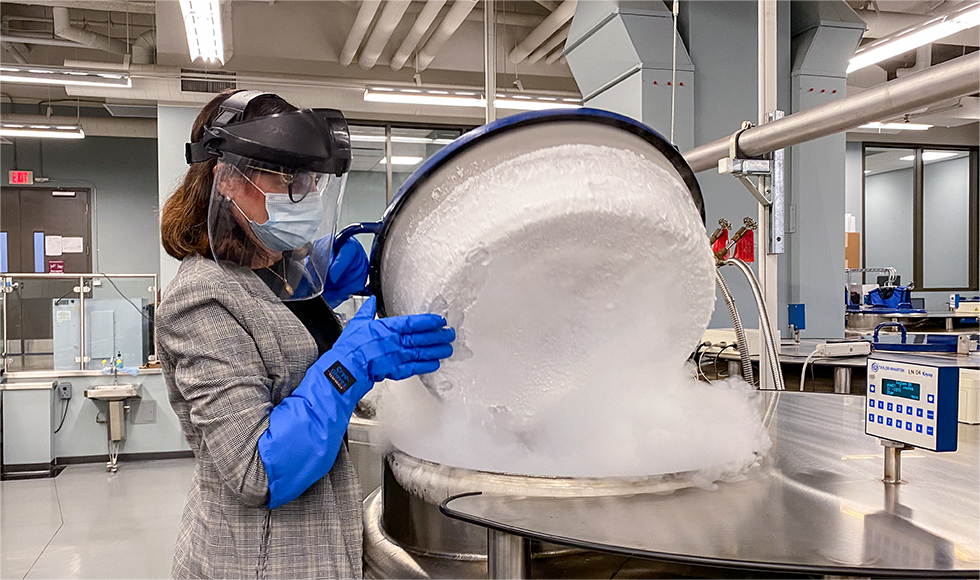 Woman in mask, face shield, protective gloves lifts up a lid to release a lot of dry-ice smoke in a lab setting.