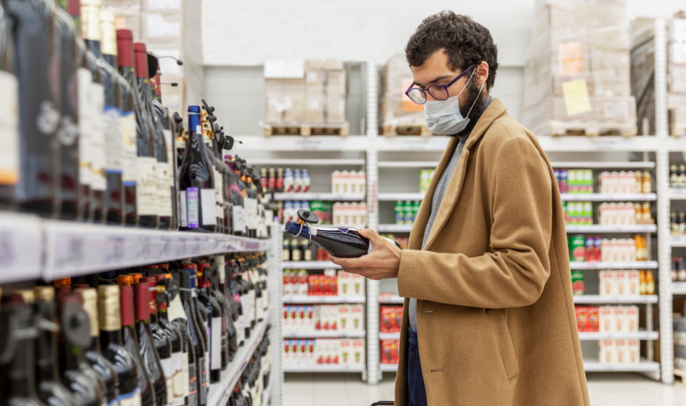 A man wearing a mask is standing in front of a shelf full of wine bottles. He is holding a bottle of wine in his left hand and is looking down at it.