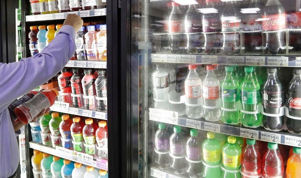 A hand reaching into a store refrigerator full of cans of cola and other pop. The full person is out of frame.