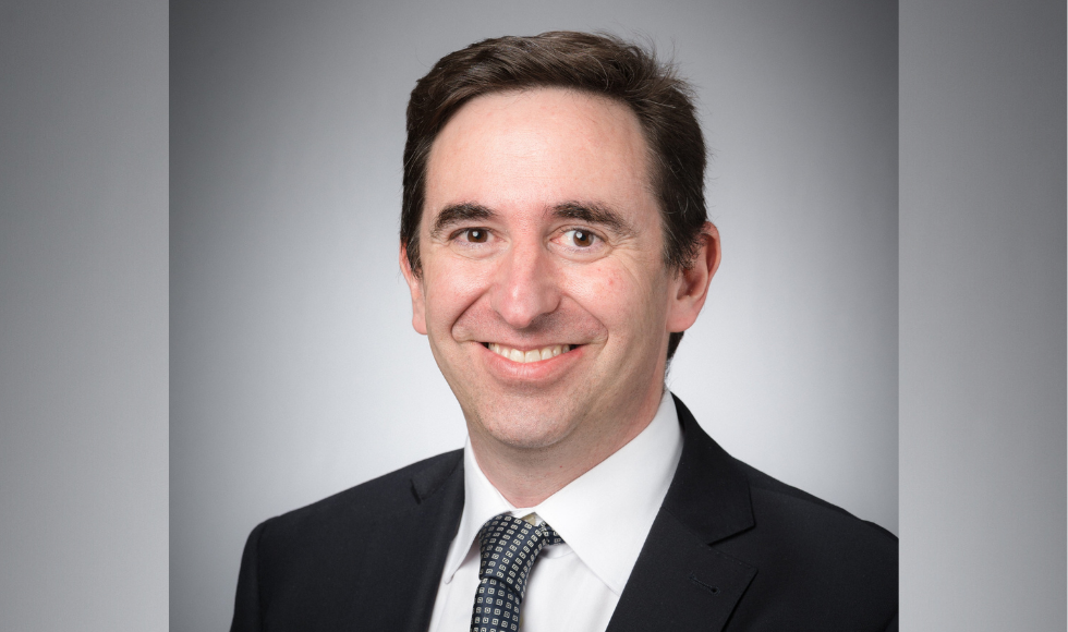 A headshot of Carlos Filipe. He is wearing a black suit jacket, white dress shirt and navy and cream tie. He is smiling at the camera and is against a grey background.