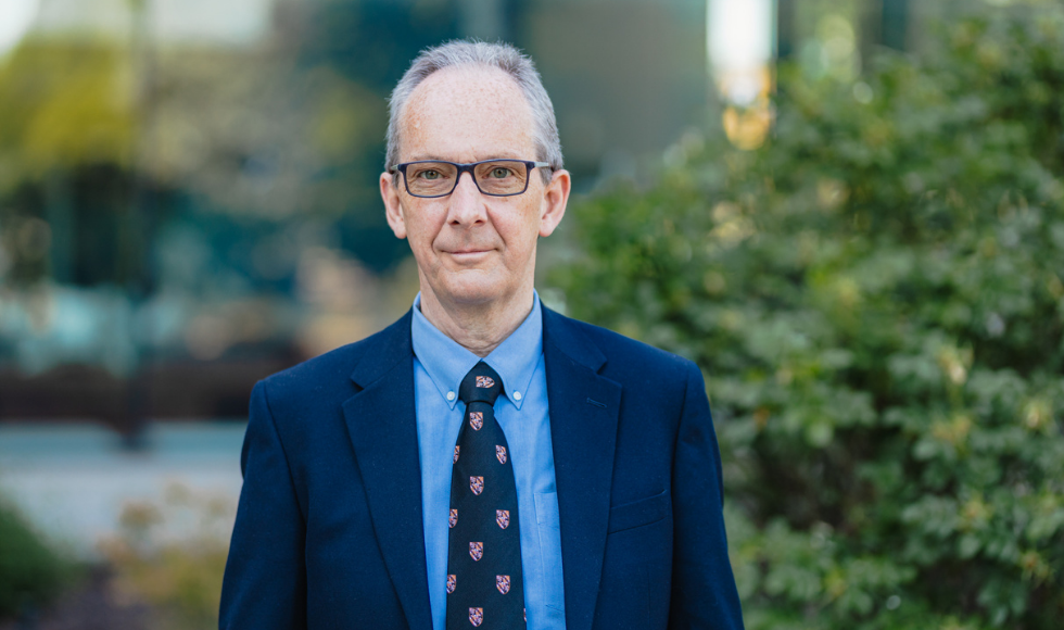 A headshot of David Armstrong, who is wearing a suit and tie. He is outside and there are bushes, trees and a building in the background.