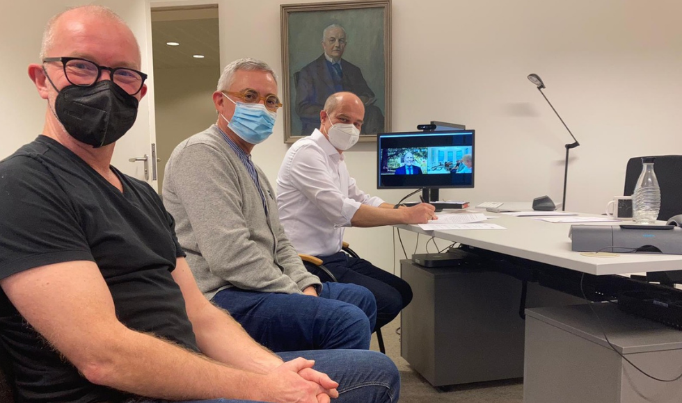 A photo of three masked men sitting in front of a desk. McMaster President David Farrar’s face is visible on a computer screen sitting on the desk. The man closest to the computer is signing a document.