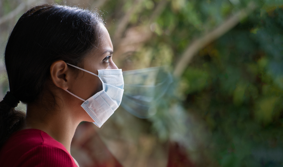 A photo of a masked woman looking out a window. Her face is not fully visible to the viewer.