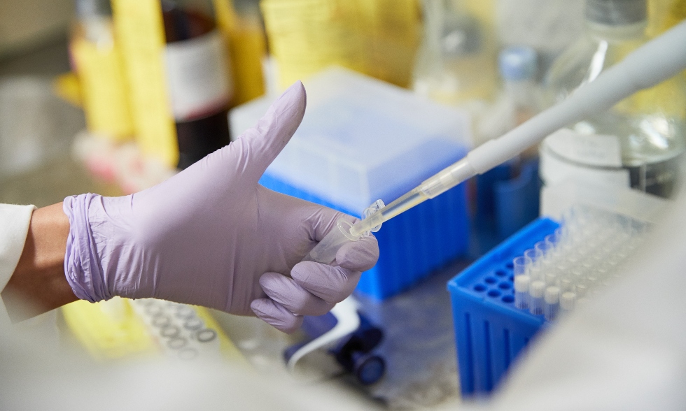 A photo of a gloved hand holding a lab instrument being filled with a liquid from a pipette. Lab instruments can be seen in the background.