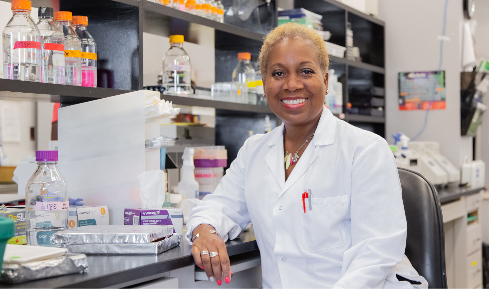 Juliet Daniel sitting at a lab desk wearing a lab coat, smiling at the camera