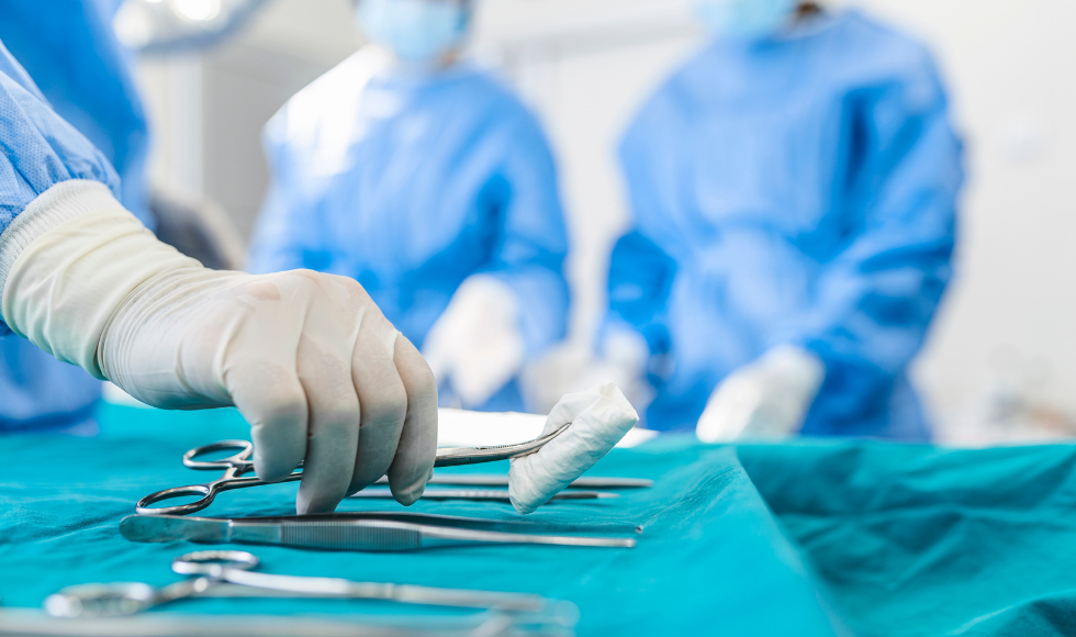 A gloved hand lifting a surgery instrument up off a surface. The bodies of two gowned surgeons can be seen in the background.