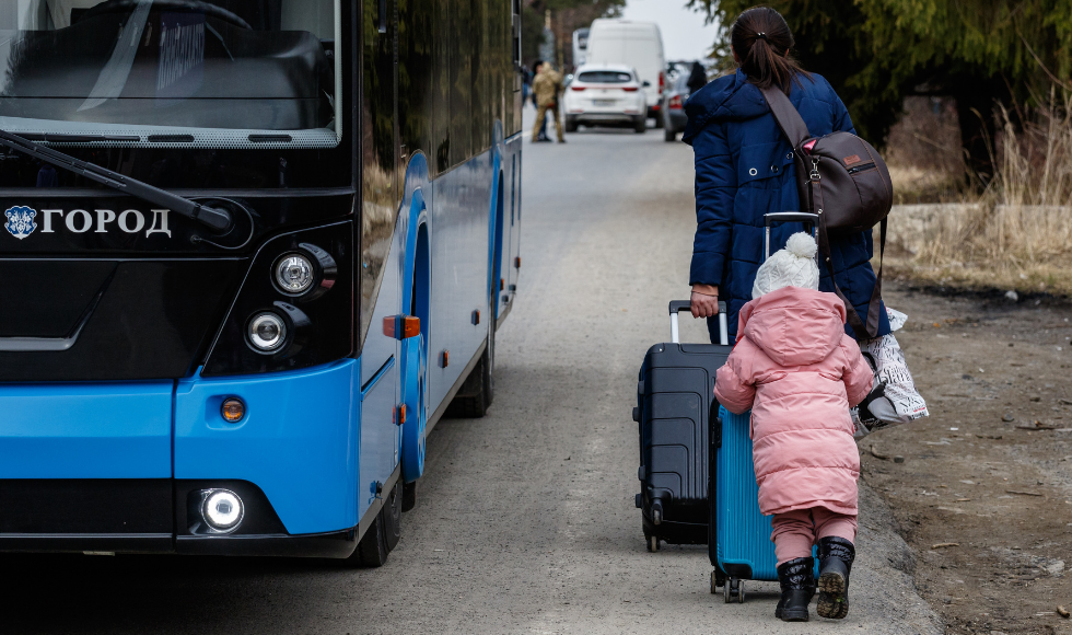 A woman pulls a suitcase on wheels along a road. A young girl in a pink coat pushes a suitcase on wheels behind her.