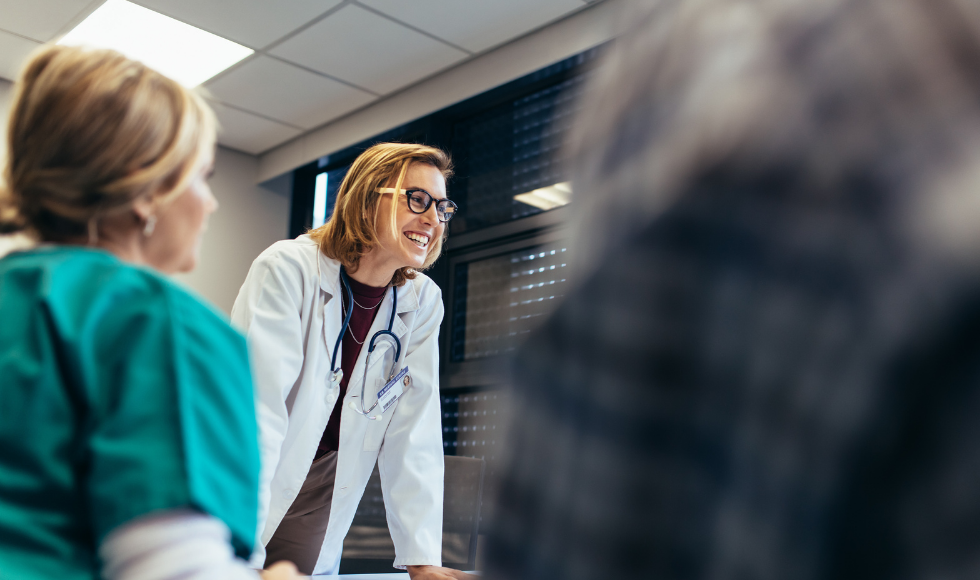 A smiling female doctor in a lab coat stands at the head of a conference table with others seated around it.