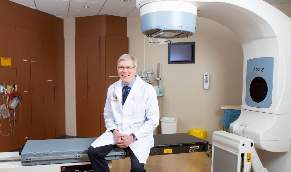 Tim Whelan wearing a lab coat, sitting in an oncology examination room.