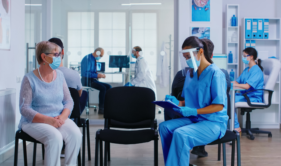 A nurse in scrubs fills out a form while talking to an older adult in a waiting room.